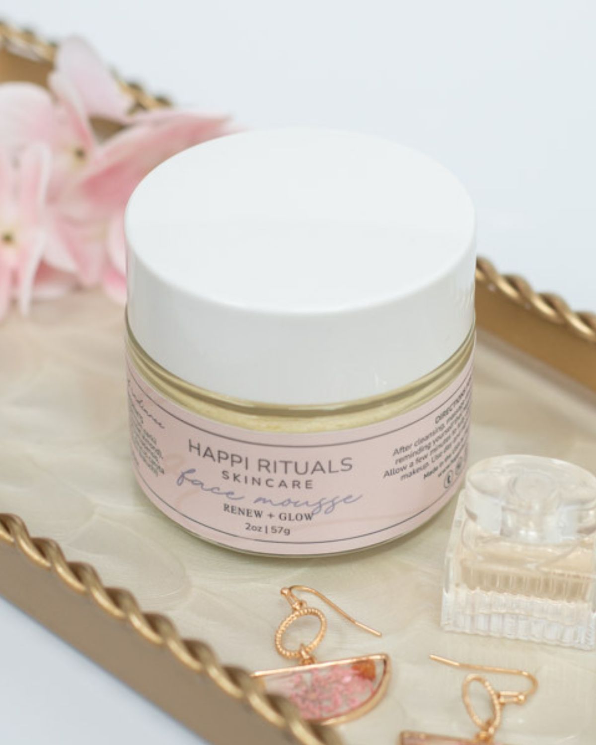 A skincare jar on a tray with jewelry and flowers, viewed from above. The jar is white with a pink label.