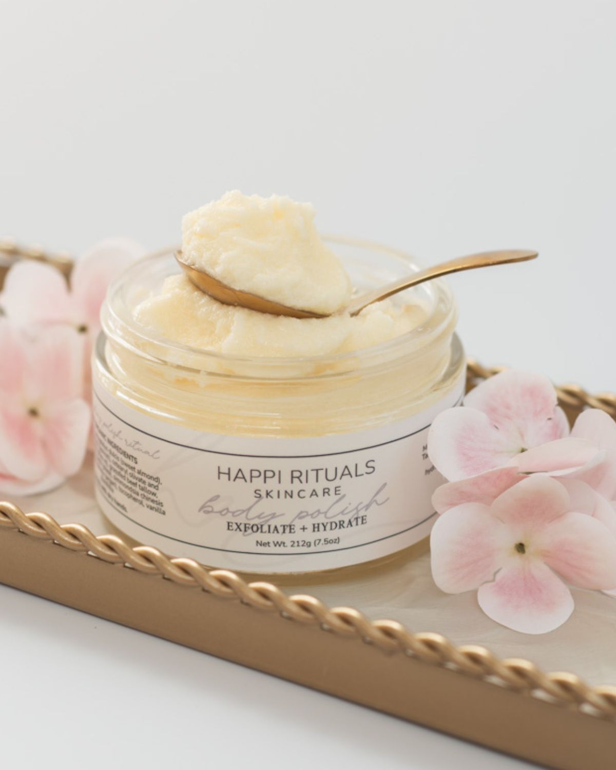 Creamy skincare product in a jar with a spoon, surrounded by pink flowers on a tray.