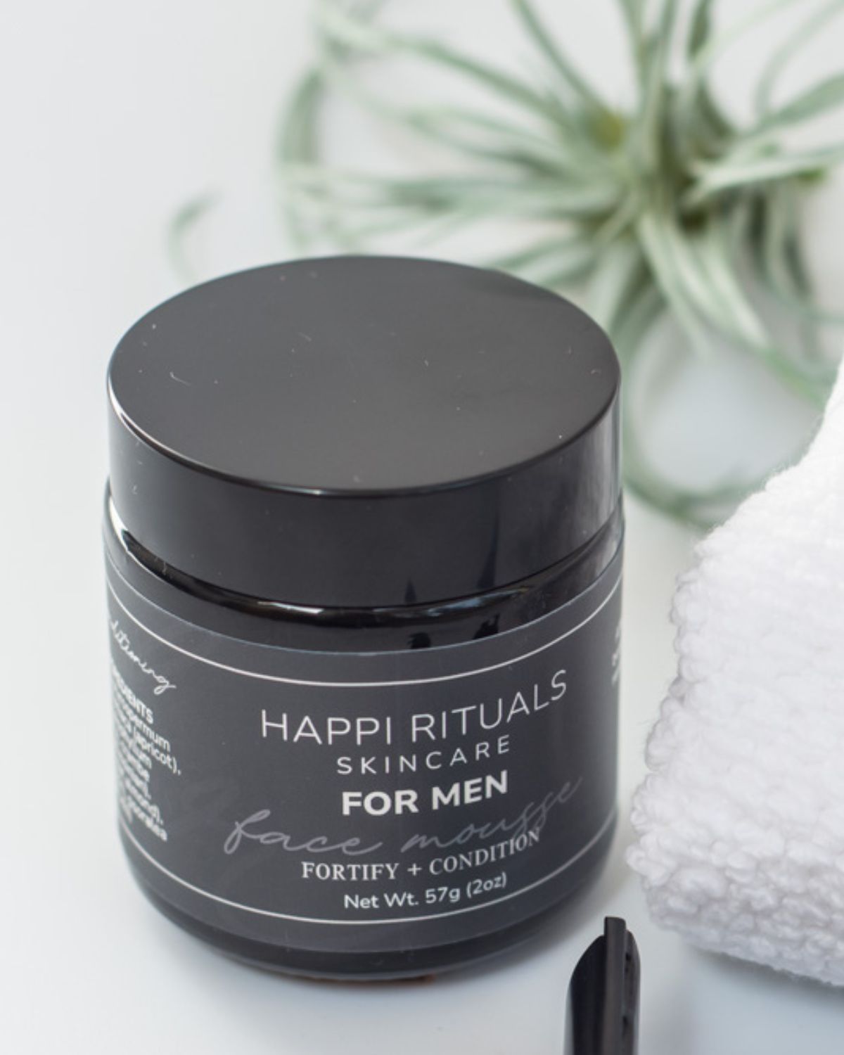 Black jar of Happi Rituals skincare for men with a white towel and plant in background.