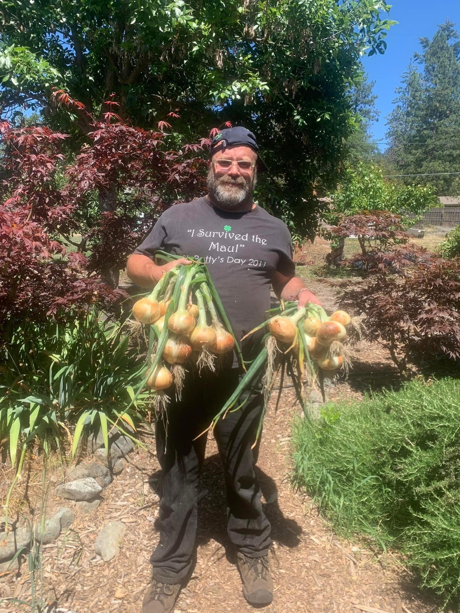 Man with a beard holding multiple onions outdoors in a garden, smiling, with trees and plants in the background.