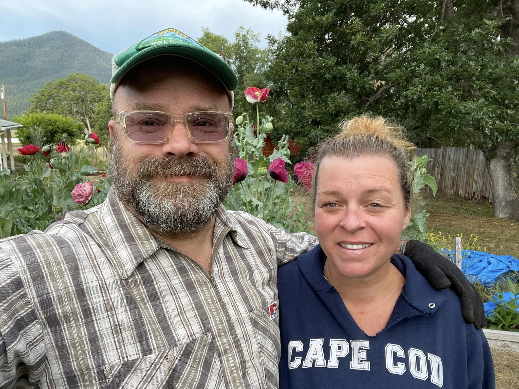 Two people smiling outdoors with flowers and trees in the background.