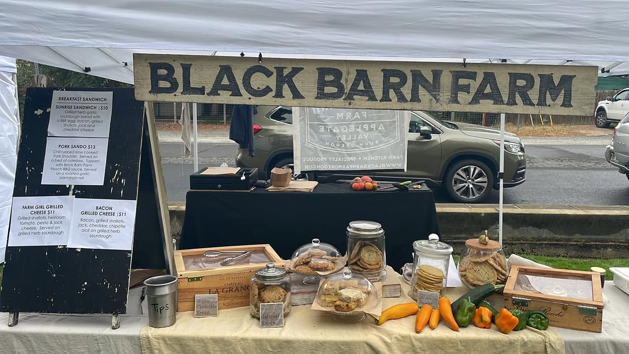 Display table with jars, vegetables, and baked goods at Black Barn Farm, with a sign overhead. Cars are visible in the background.