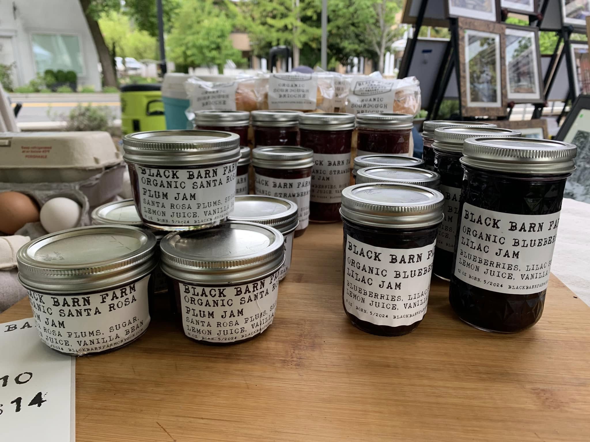 Multiple jars of organic jam on a wooden table, with framed pictures and greenery in the background.