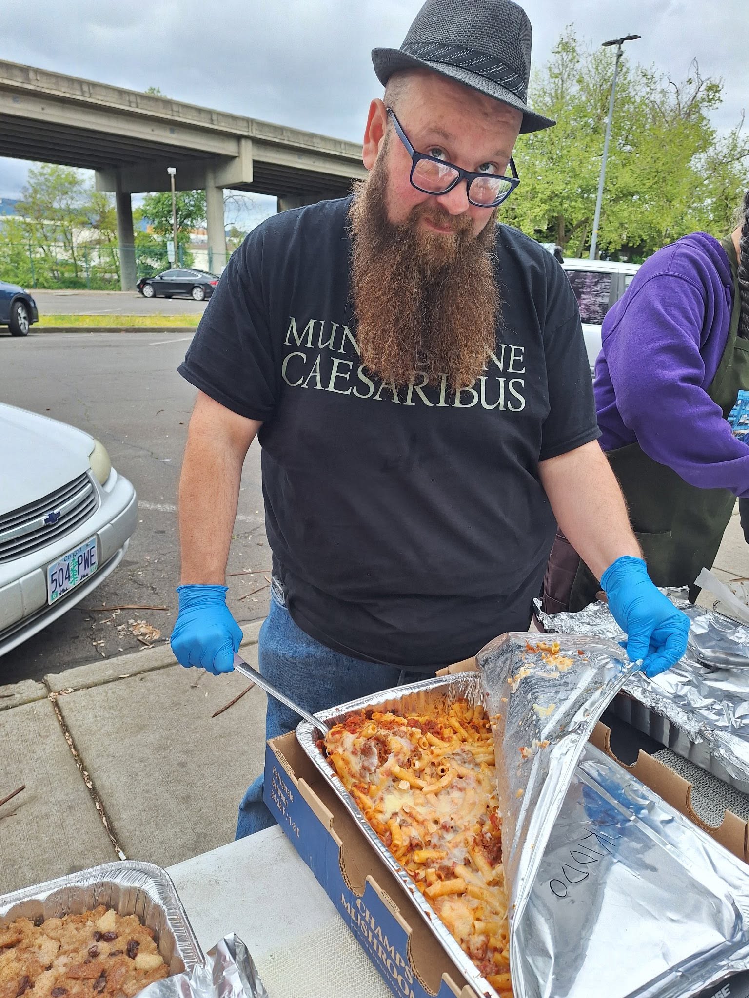 Man with glasses, beard, and hat serving pizza outdoors, wearing blue gloves, under a bridge, with trees and cars nearby.