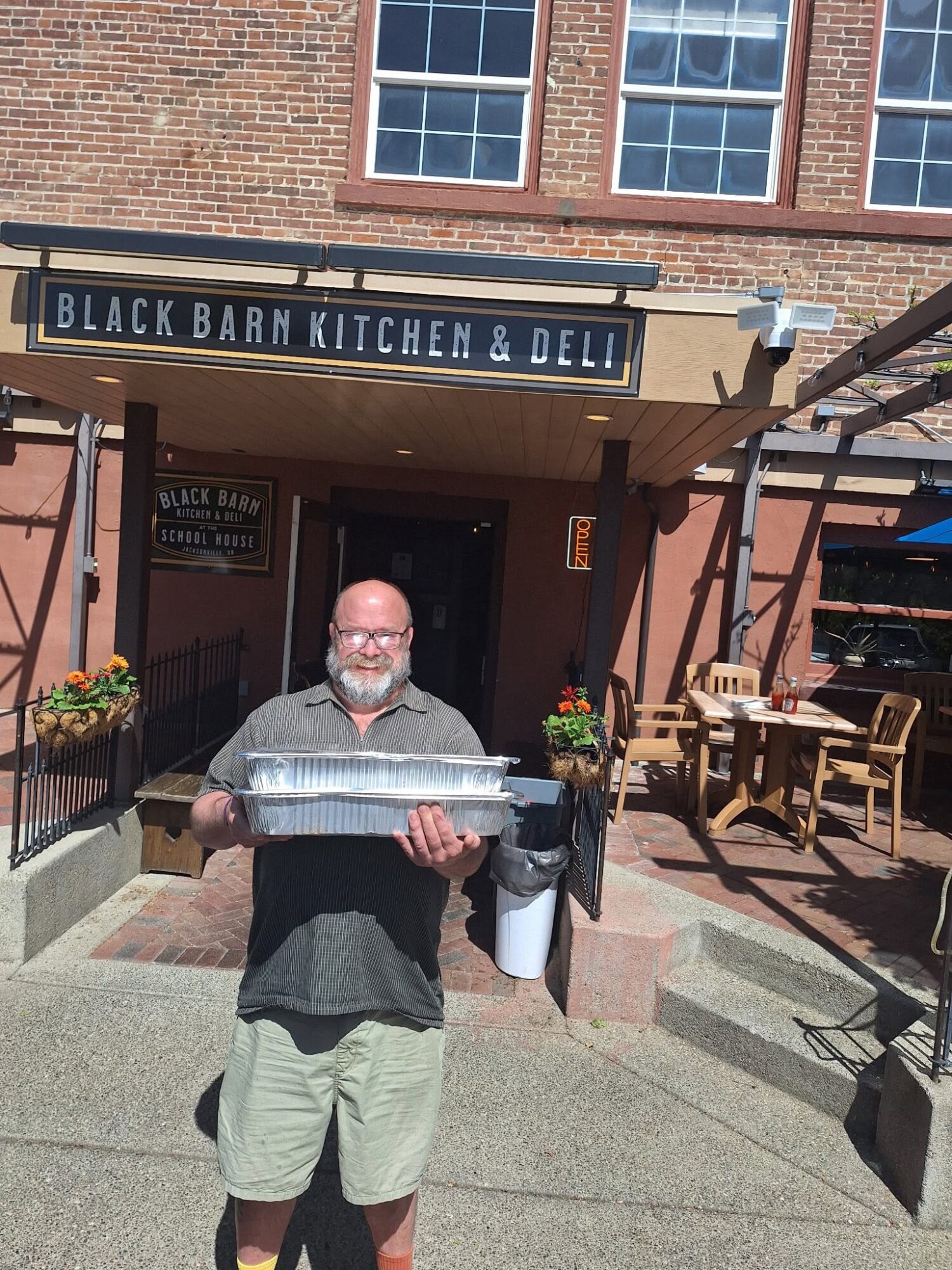 Man with glasses and beard holding a large fish outside a restaurant with a sign reading 'Black Barn Kitchen & Deli'.
