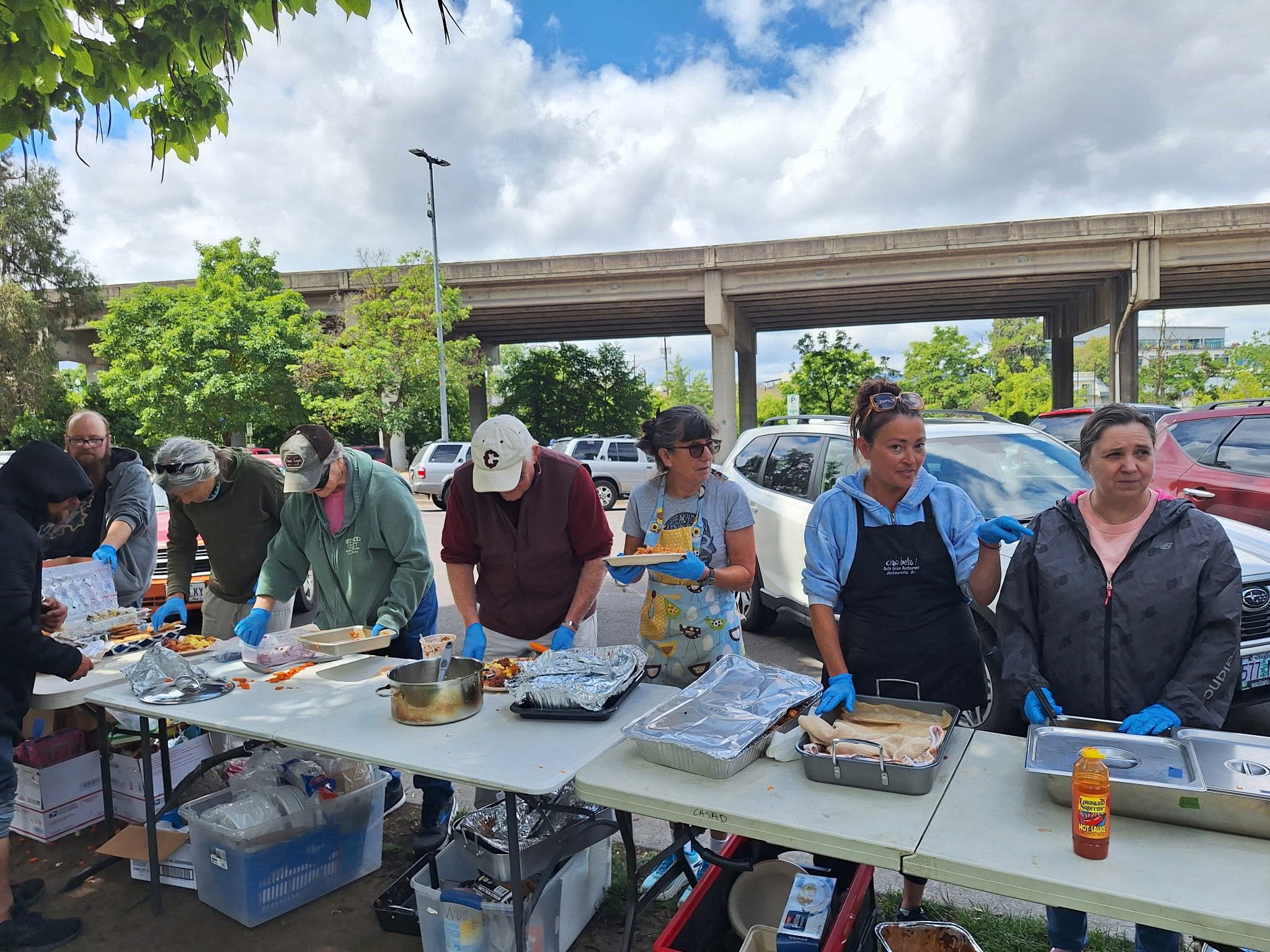 People preparing food at an outdoor table with a bridge and cars in the background, under a partly cloudy sky.