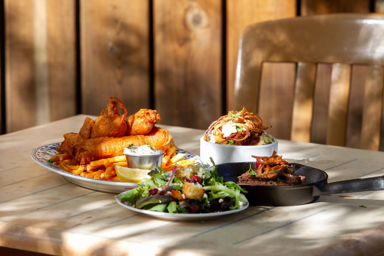Three plates of food on a table with a wooden wall background, including fried chicken, fries, salad, and a burger.