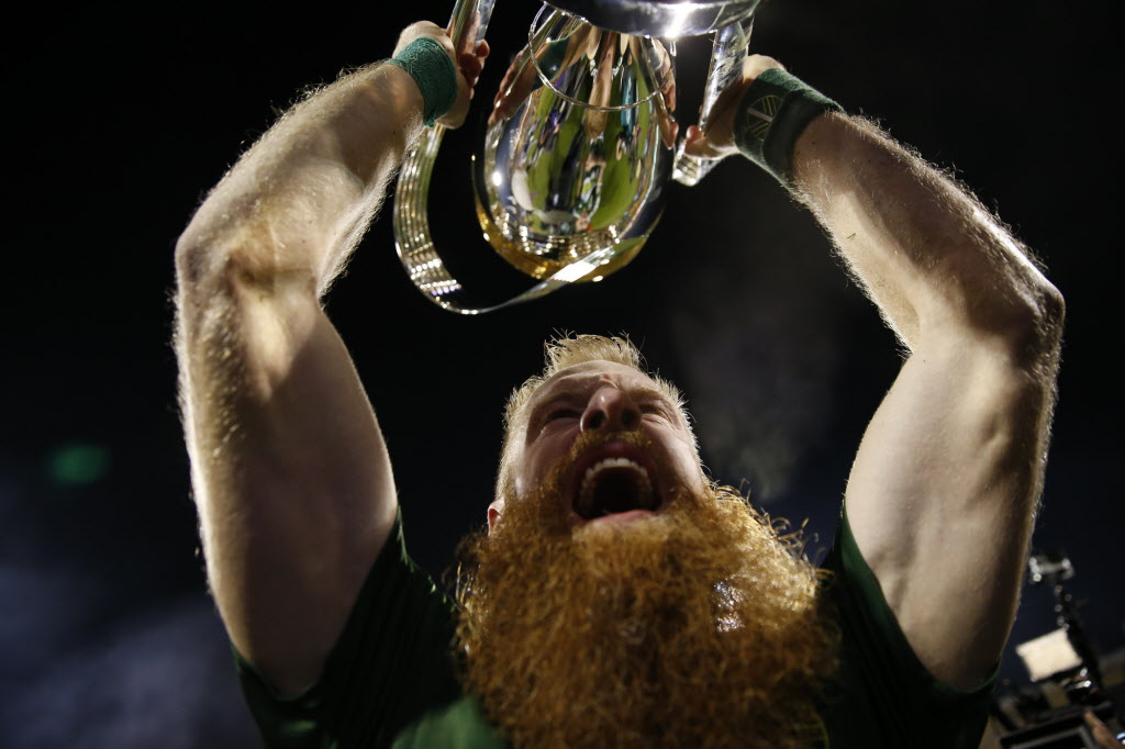 A bearded man with arms raised holding a trophy, celebrating victory, with a dark background.