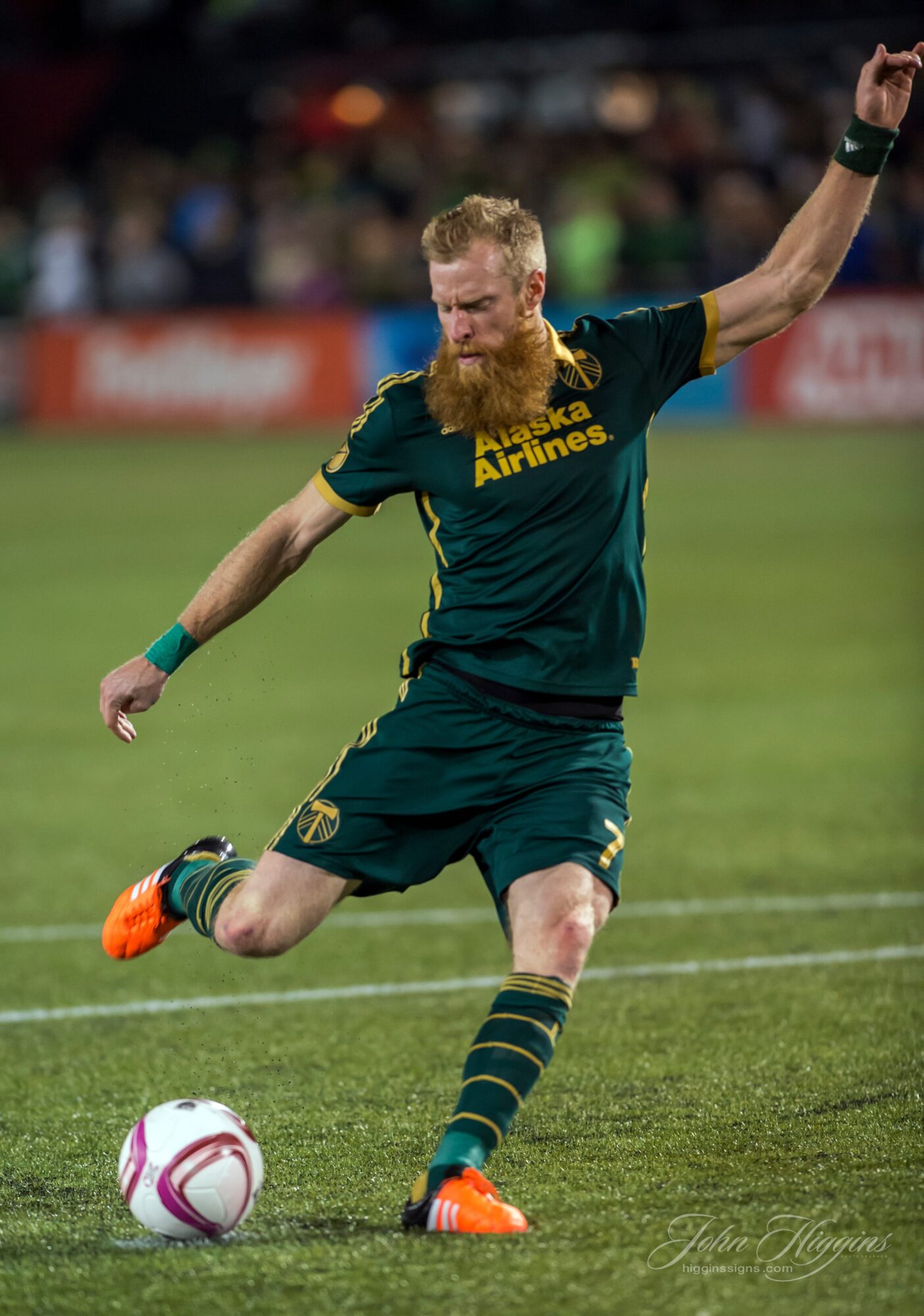 A soccer player with a beard and long hair kicks a ball on the field, wearing a dark green uniform with yellow accents.