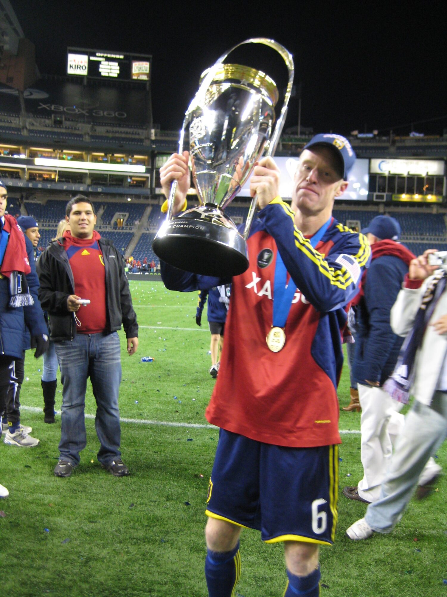 Person in sports uniform holding a large trophy, celebrating on a field with other people in the background.
