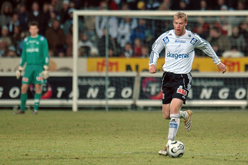 Soccer player in white jersey kicking ball on field with goalkeeper in green in background
