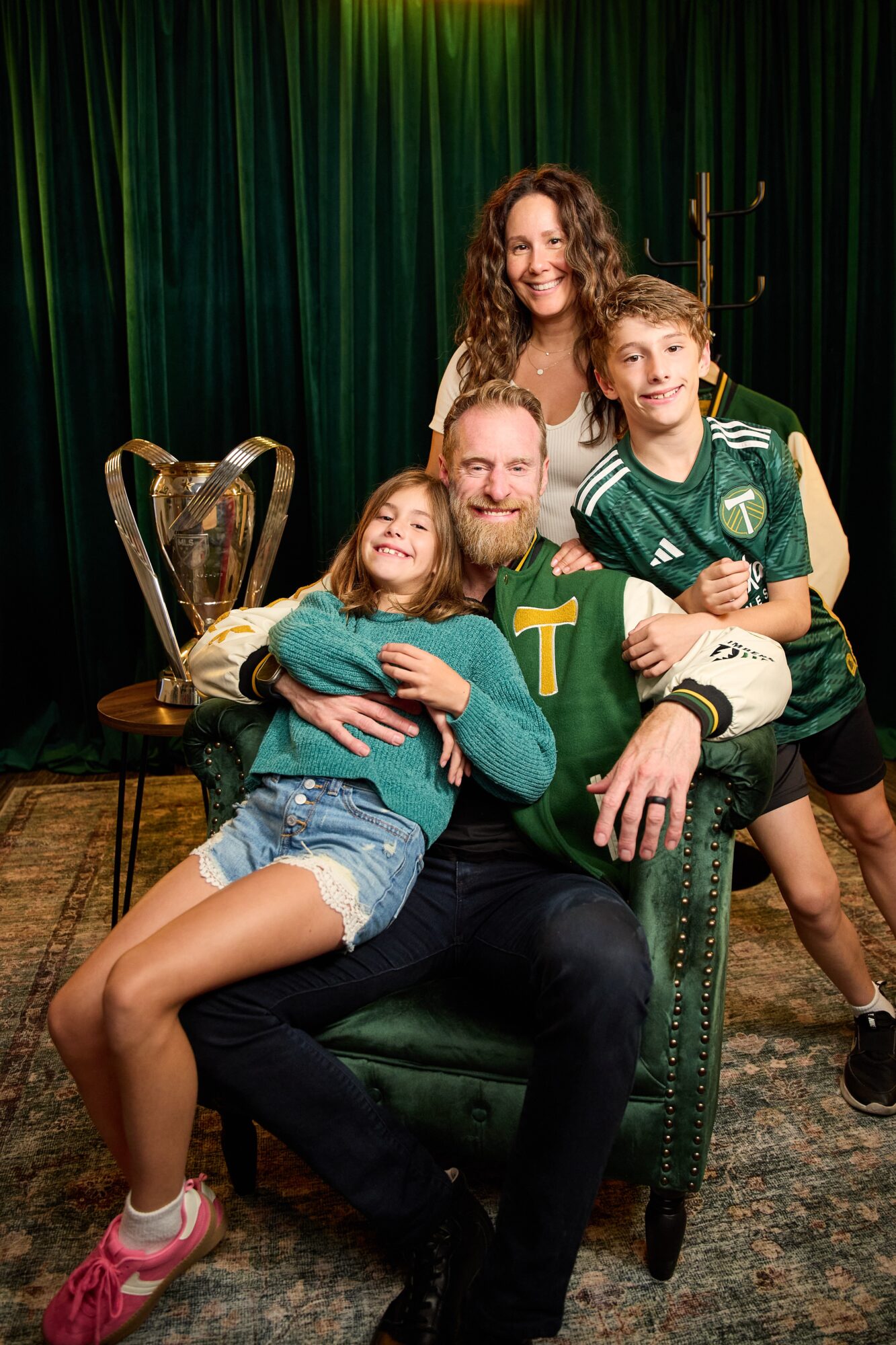 Family of four with a trophy, sitting and standing in front of green curtains, smiling, in a cozy room.