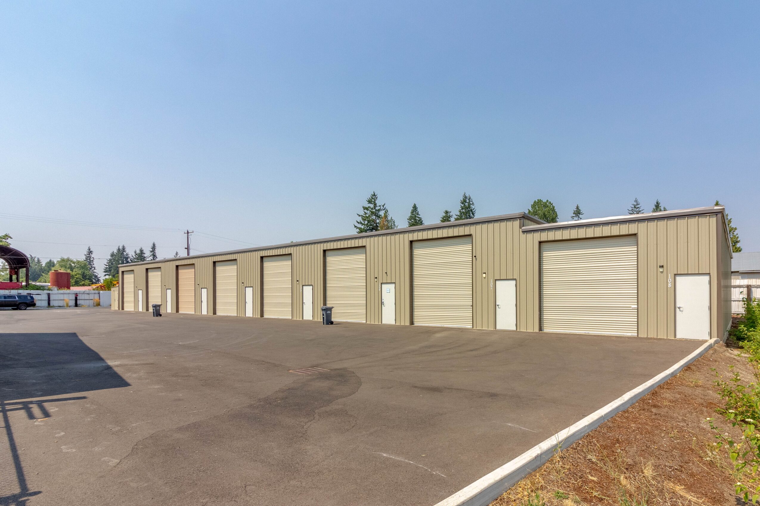 Long beige storage units with roll-up doors in a row, on a paved lot under a clear blue sky.