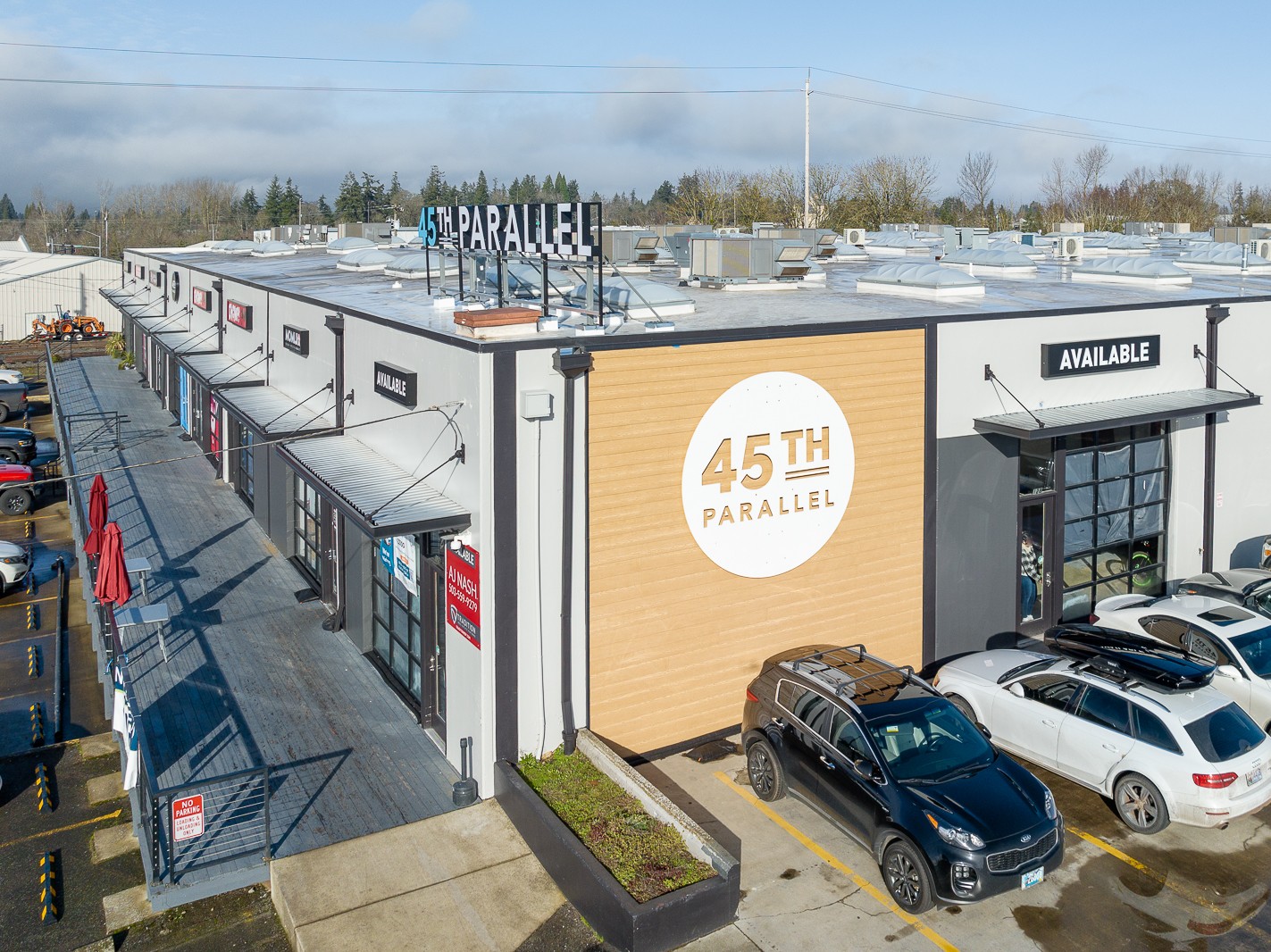 Shopping center with storefronts, parking lot, and cars, under a partly cloudy sky.