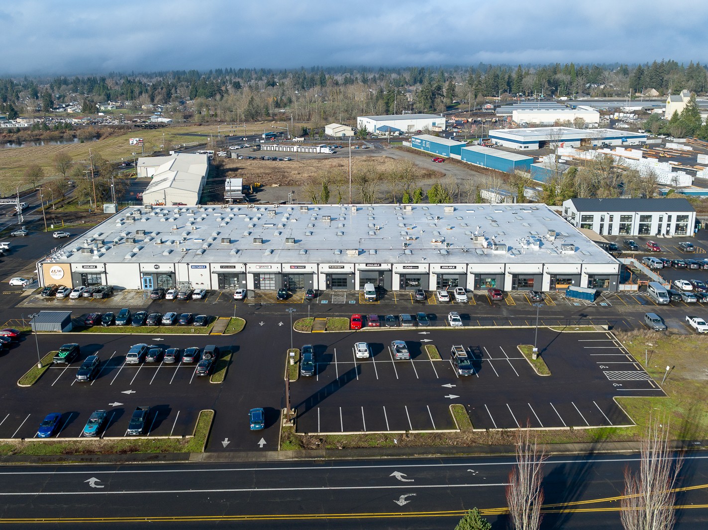 Large commercial building with parking lot in front, surrounded by trees and industrial area, under cloudy sky.