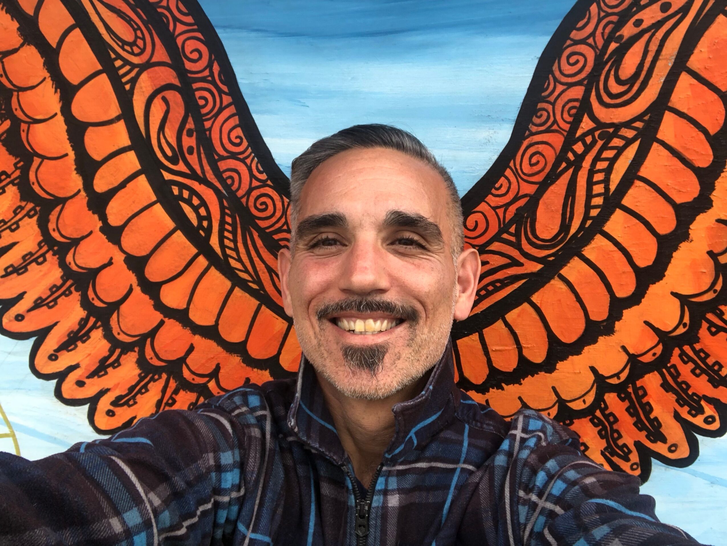 Person with sunglasses smiling, upside down, in front of orange and black patterned background, blue sky visible.