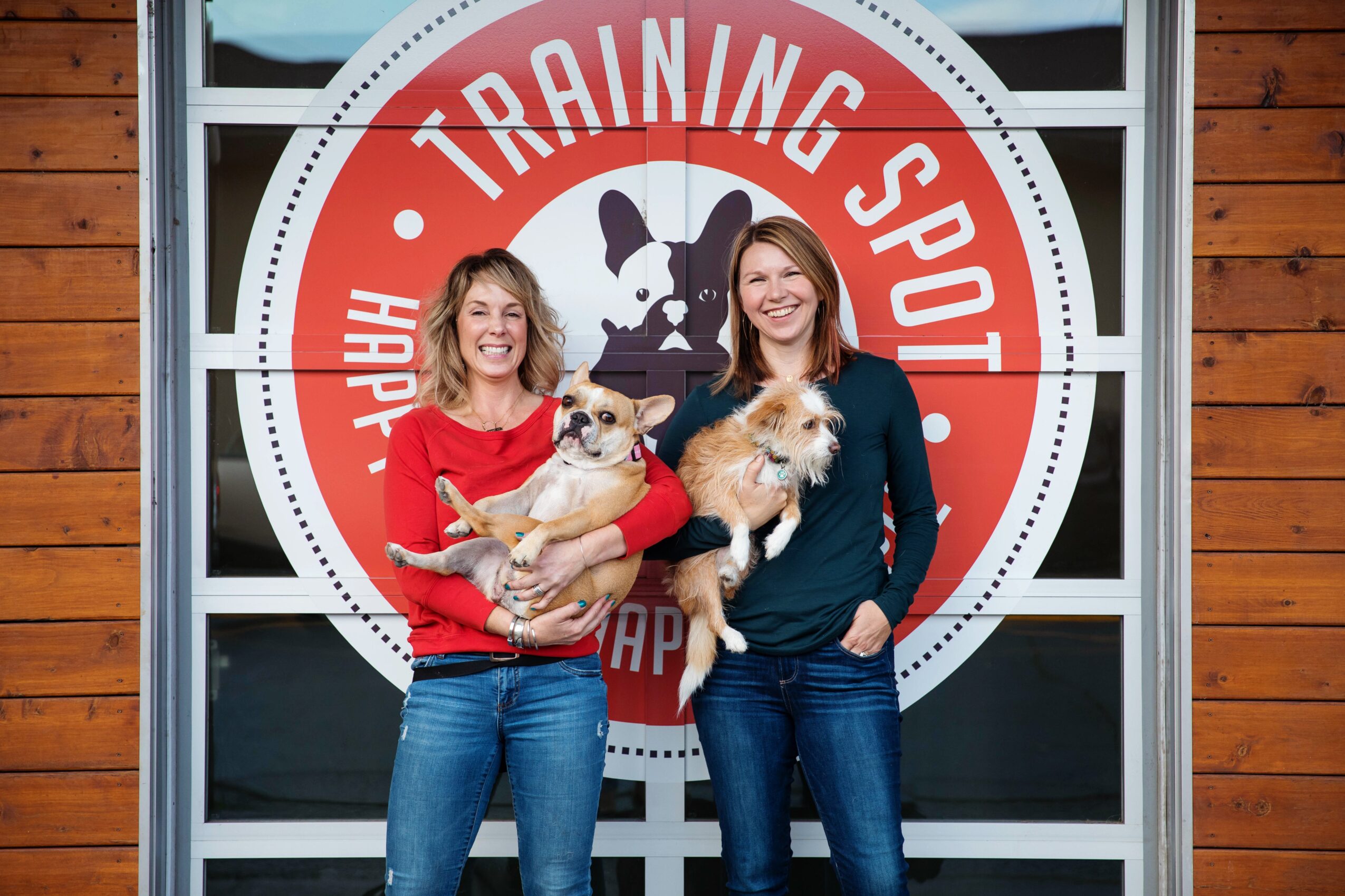 Two women holding small dogs in front of a training sports sign with a dog silhouette, wooden wall background.