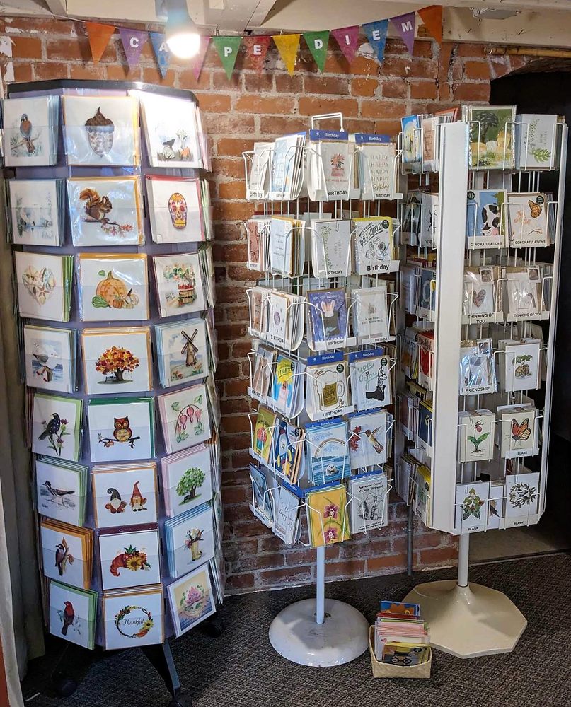 Display of greeting cards on three rotating stands against a brick wall with colorful banner overhead.