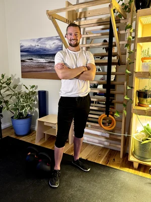 Man with beard and white t-shirt standing with arms crossed in a room with plants and a staircase.