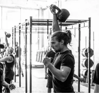 Person lifting a kettlebell overhead in a gym with others exercising nearby.