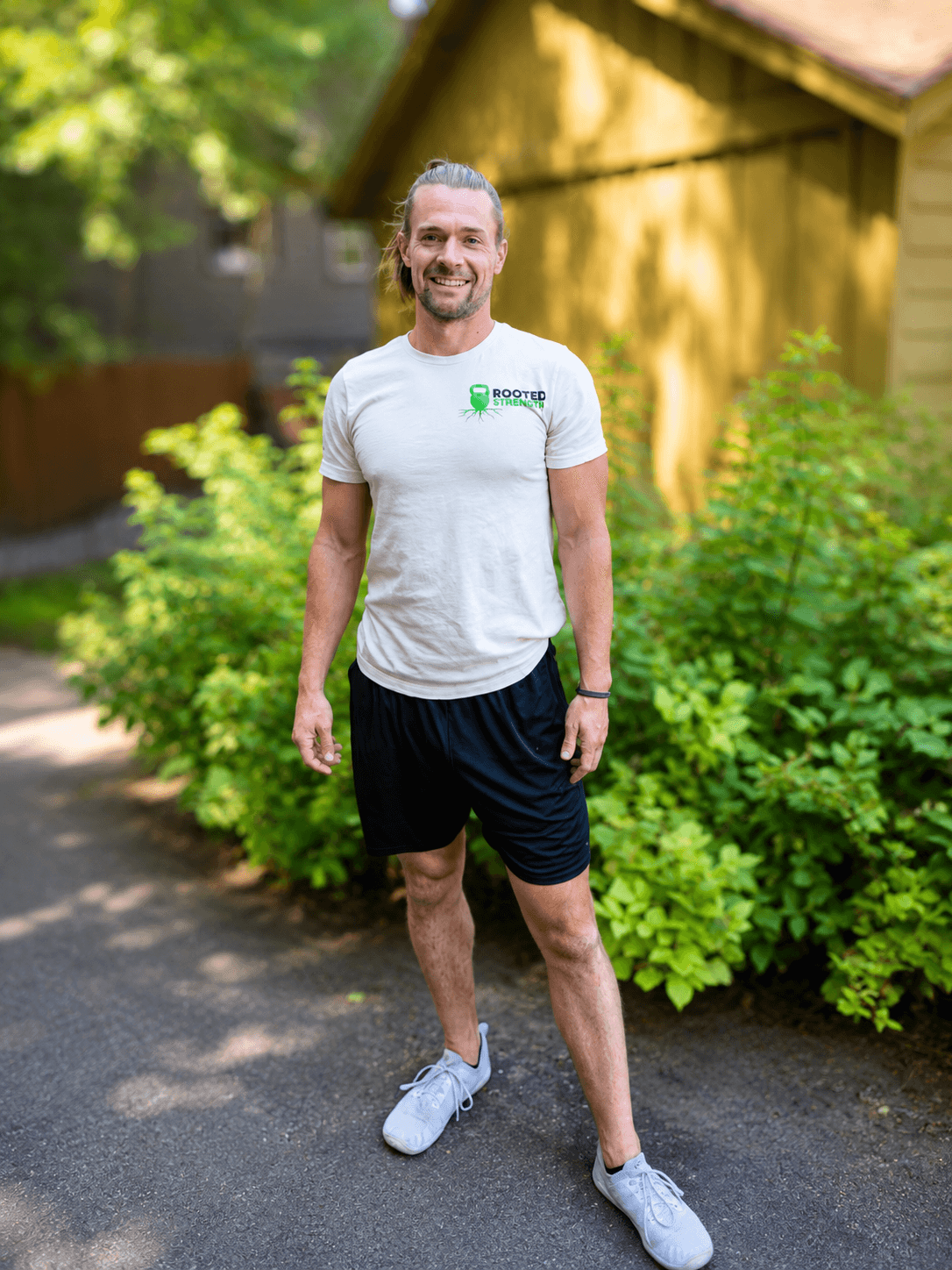 Man standing outdoors on a paved path, smiling, wearing a white t-shirt, black shorts, and white sneakers, with greenery and a wooden structure in background.