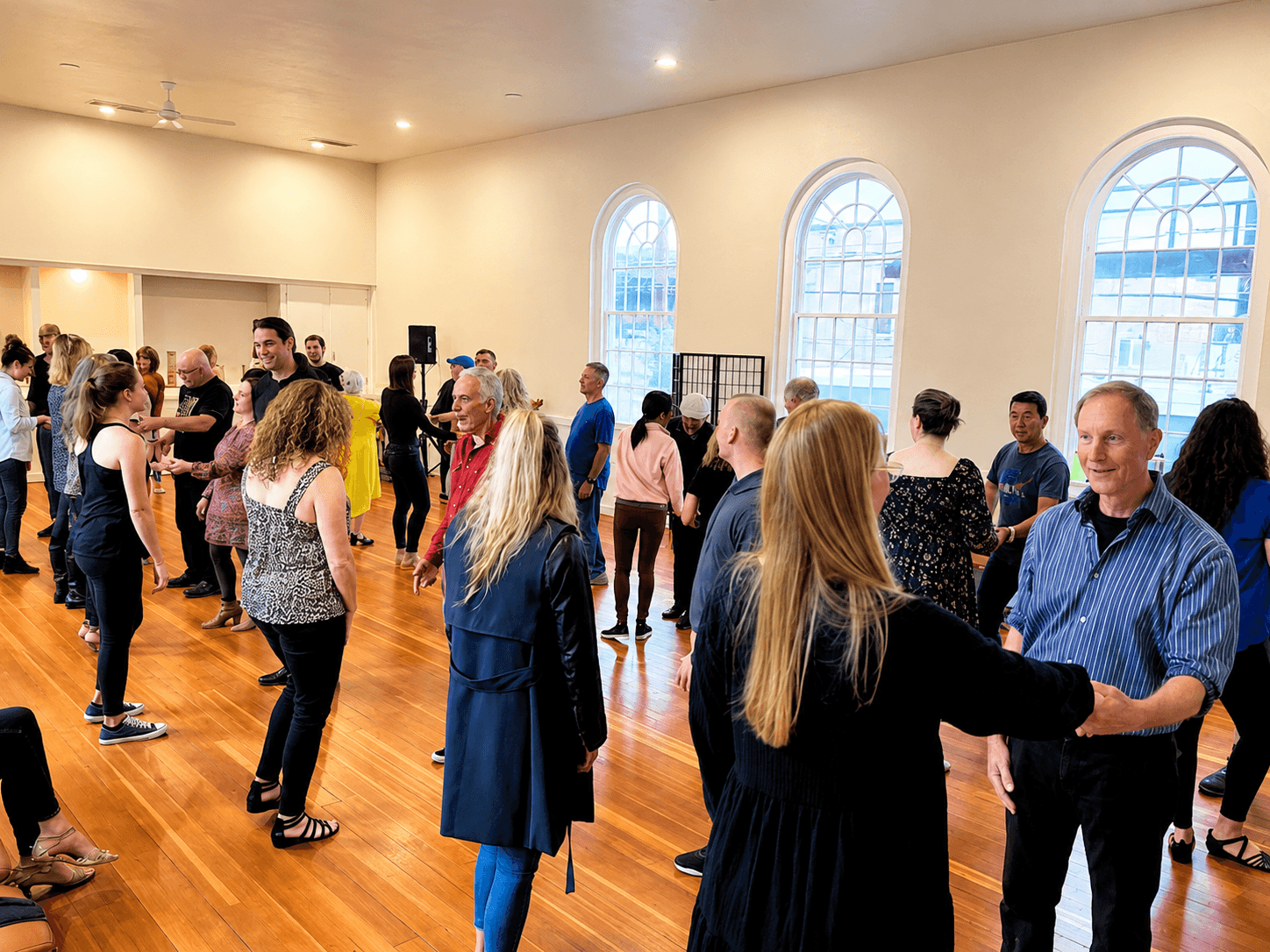 People dancing in a spacious room with large arched windows and wooden floors, some holding hands and smiling.