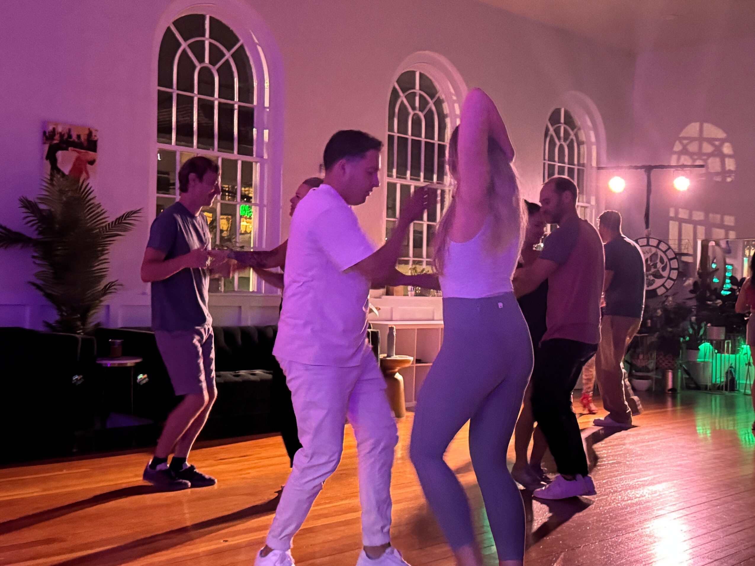People dancing in a spacious room with large arched windows and wooden floors, illuminated by colorful lights.