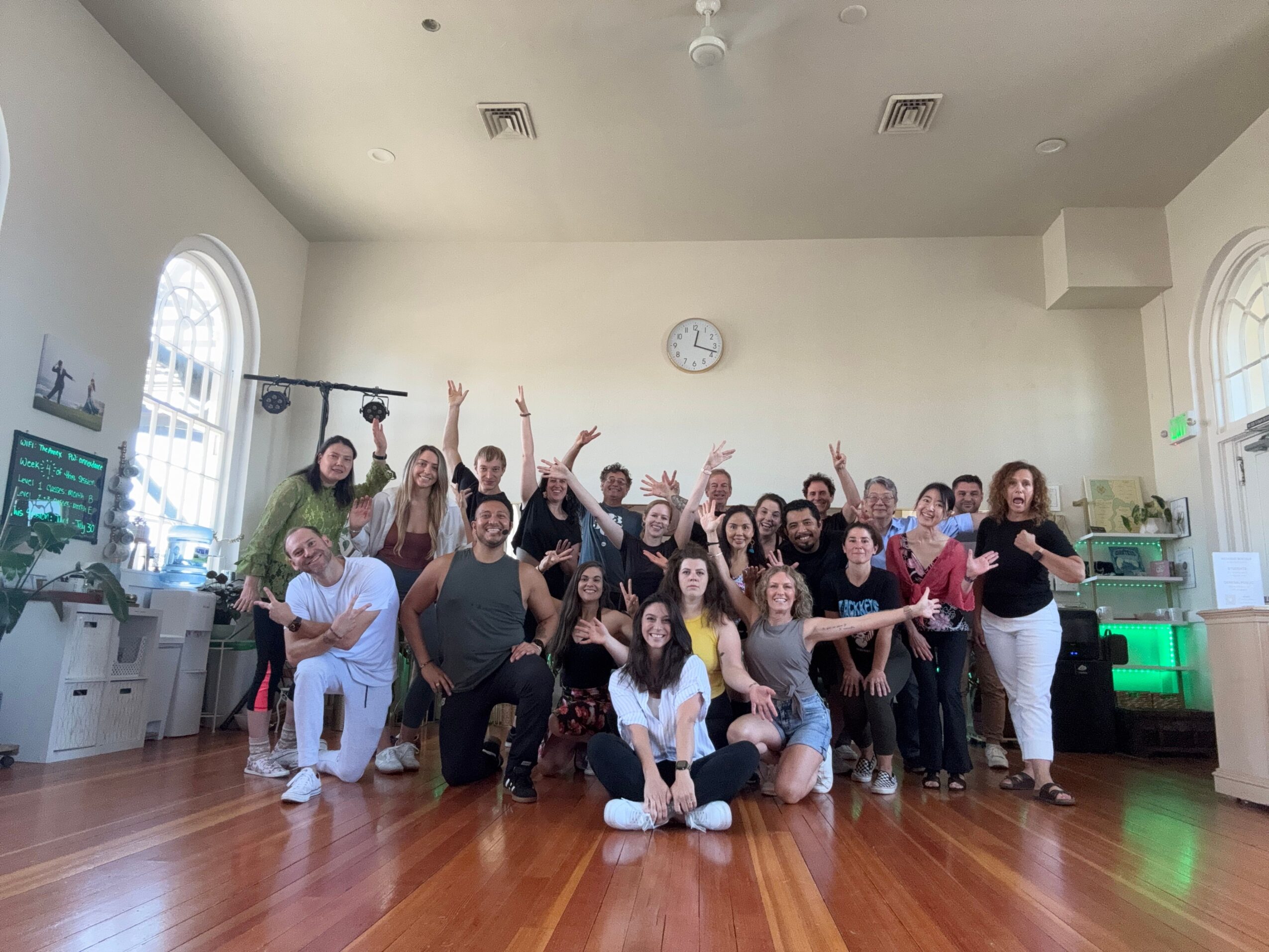 Group of people gathered in a room with wooden floor, white walls, and large windows, some raising hands, some smiling.
