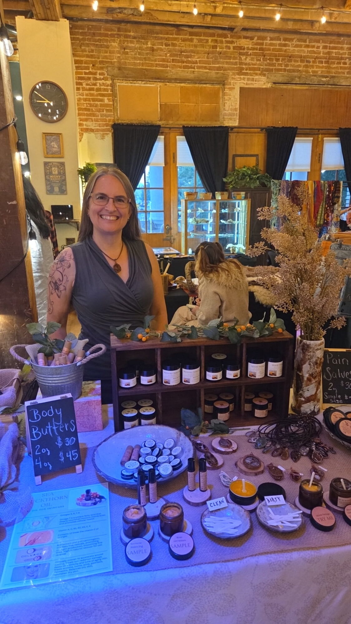 Woman smiling behind a display table with jars and baked goods, in a decorated indoor setting with brick walls and windows.
