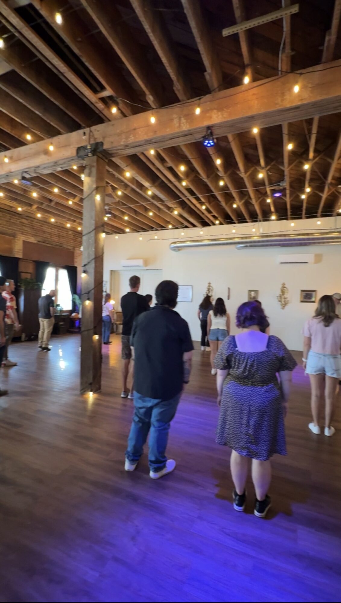 People gather in a spacious indoor venue with wooden beams and string lights overhead, some facing a white wall or screen.
