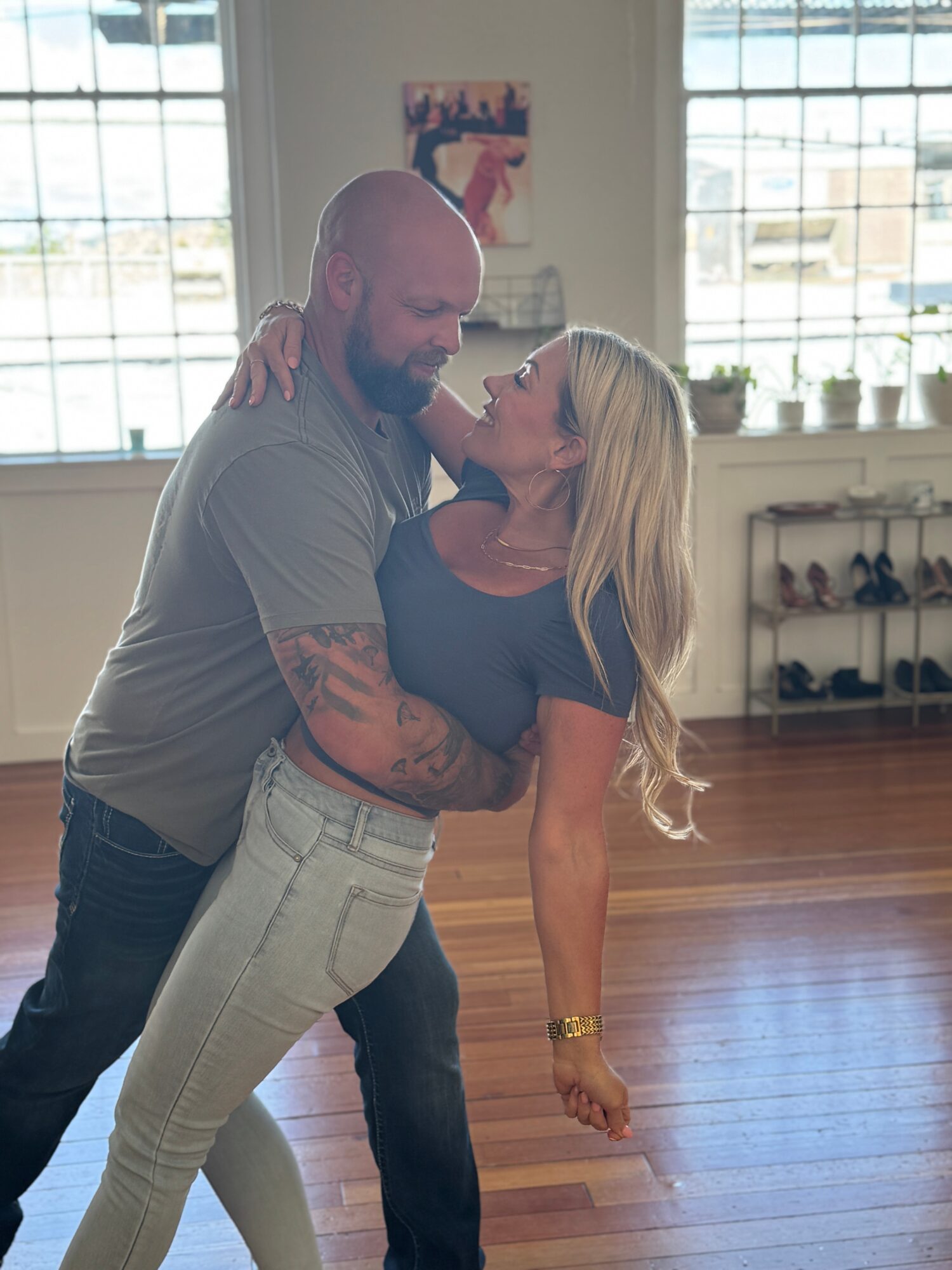 A man and woman dance closely indoors with large windows and wooden floors, smiling at each other.