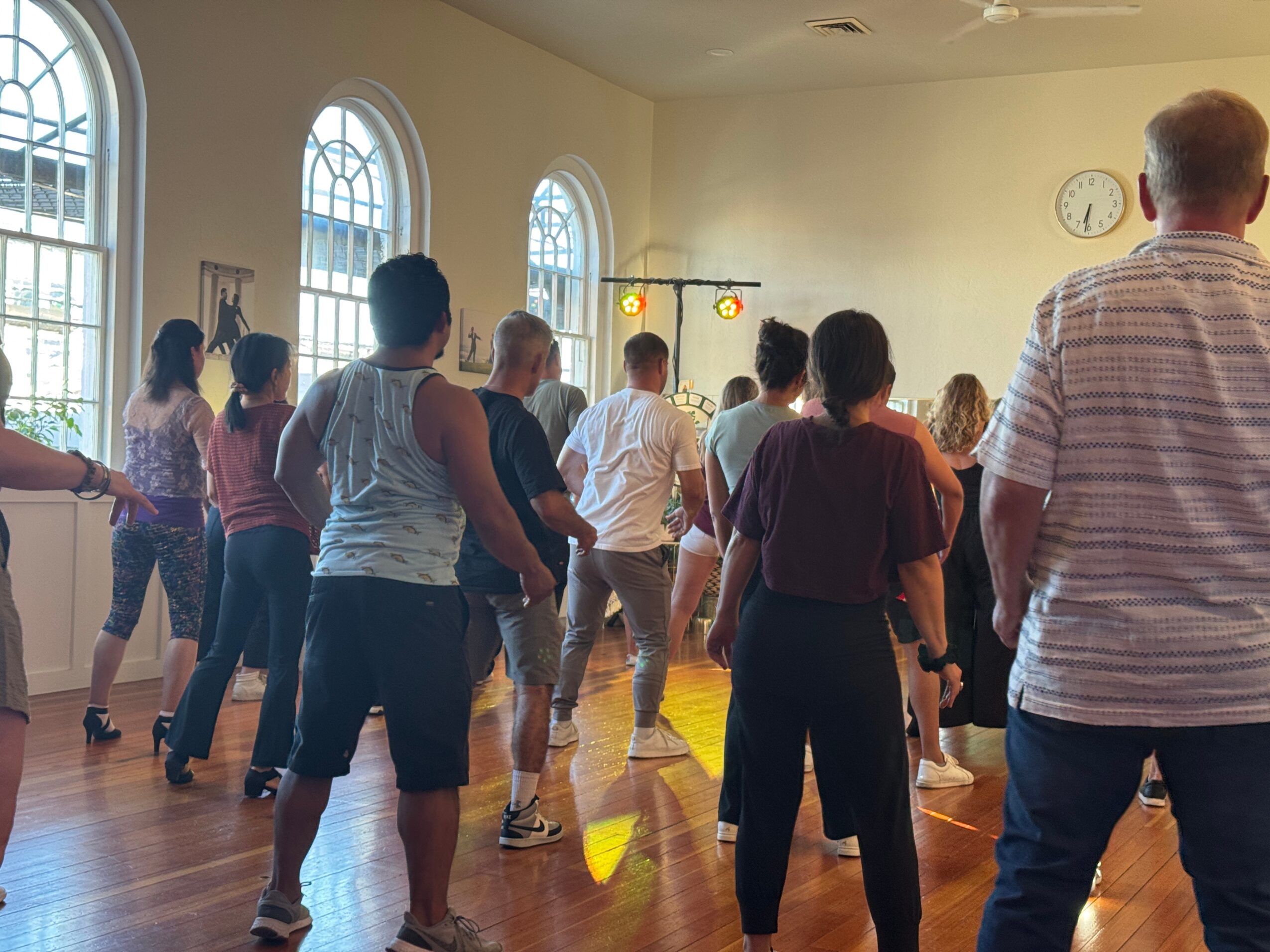 People dancing in a spacious room with large arched windows and a clock on the wall.