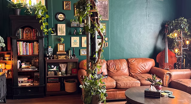 Living room with a brown leather sofa, green wall, bookshelf, plants, and decorative items.
