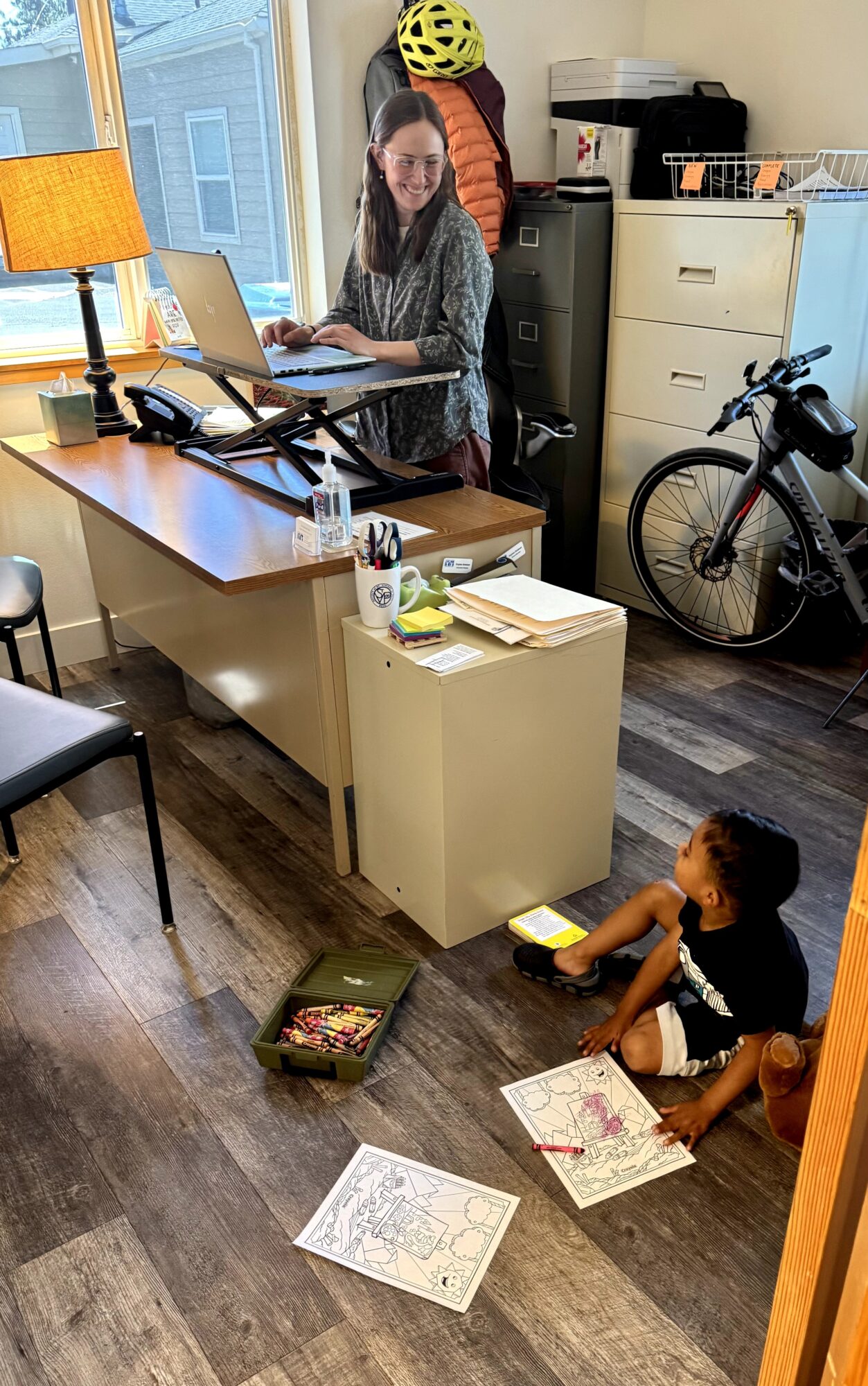 Woman working on laptop at desk, child sitting on floor with coloring books, bicycle near cabinet, window with view outside, warm lighting.