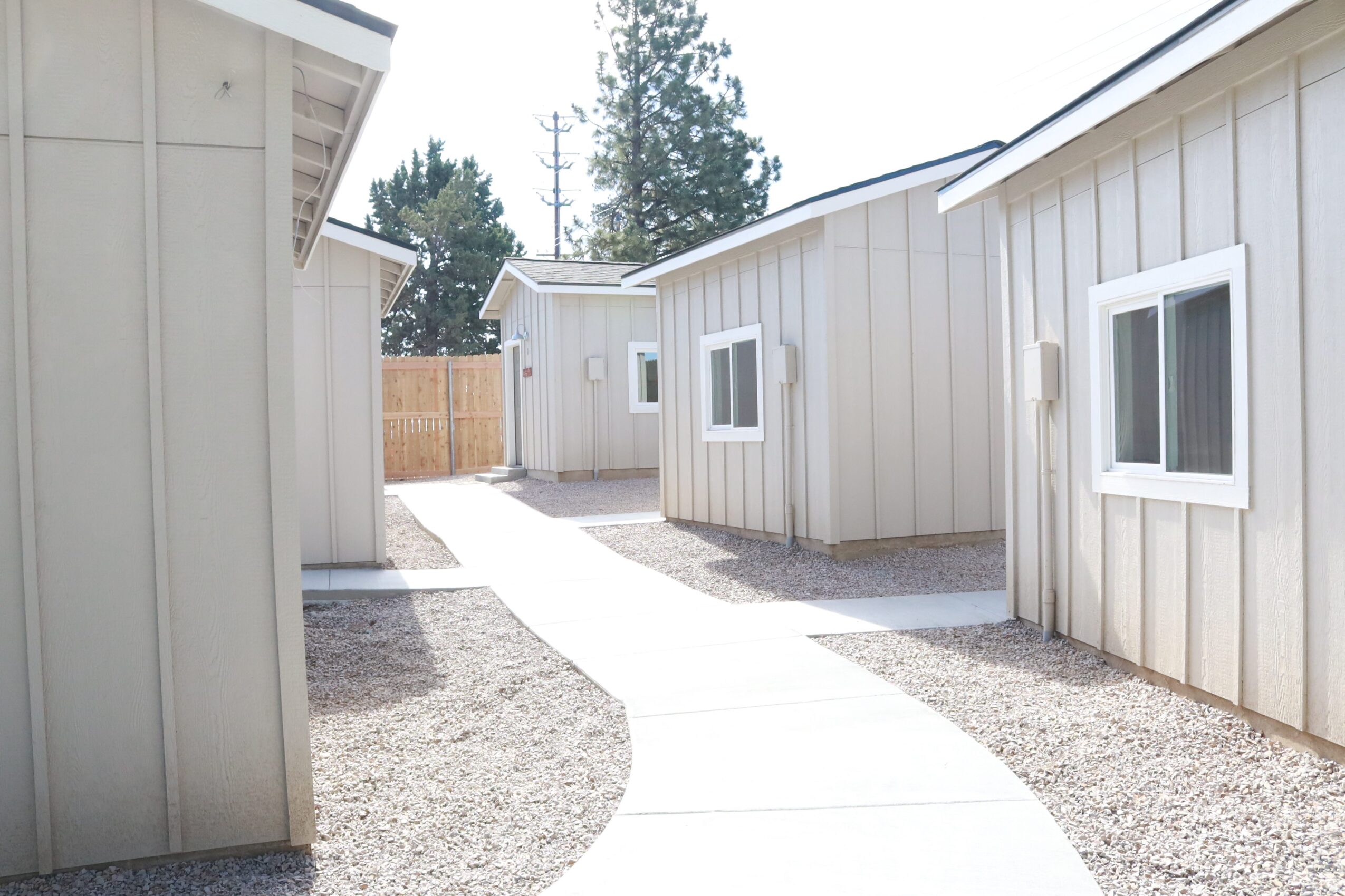 Pathway between small white buildings with windows, leading to a wooden fence, under a clear sky.