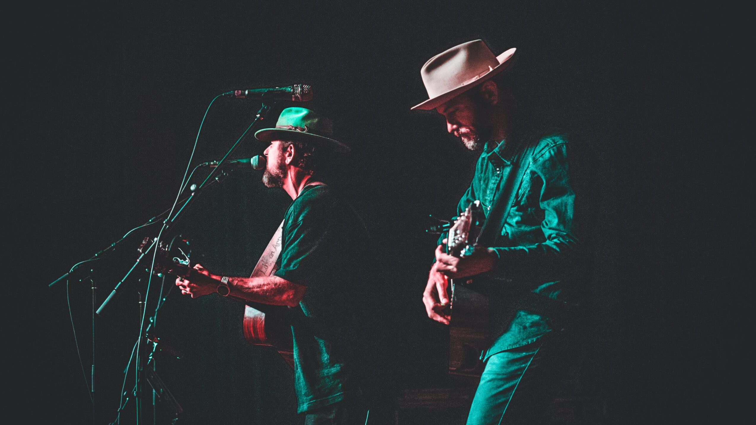 Two musicians perform on stage, one with a hat and guitar, the other with a hat and microphone, in a dark setting.