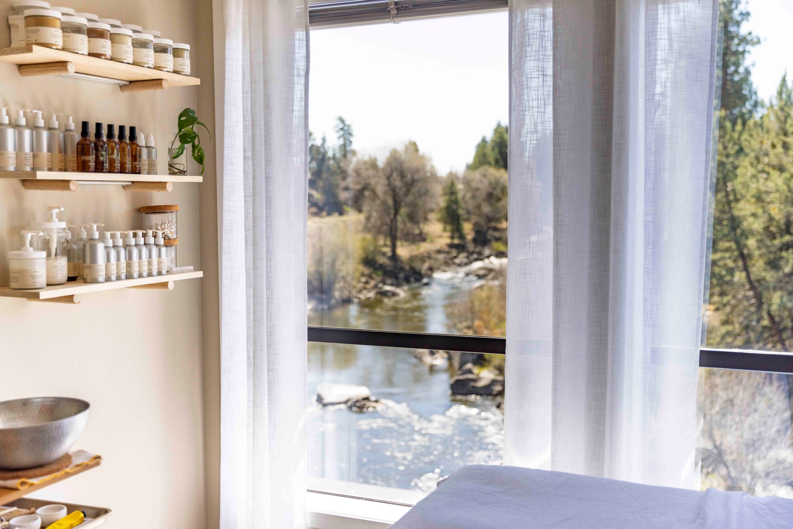 View of a river through a window with white curtains, with shelves of dishes and bowls inside a room.