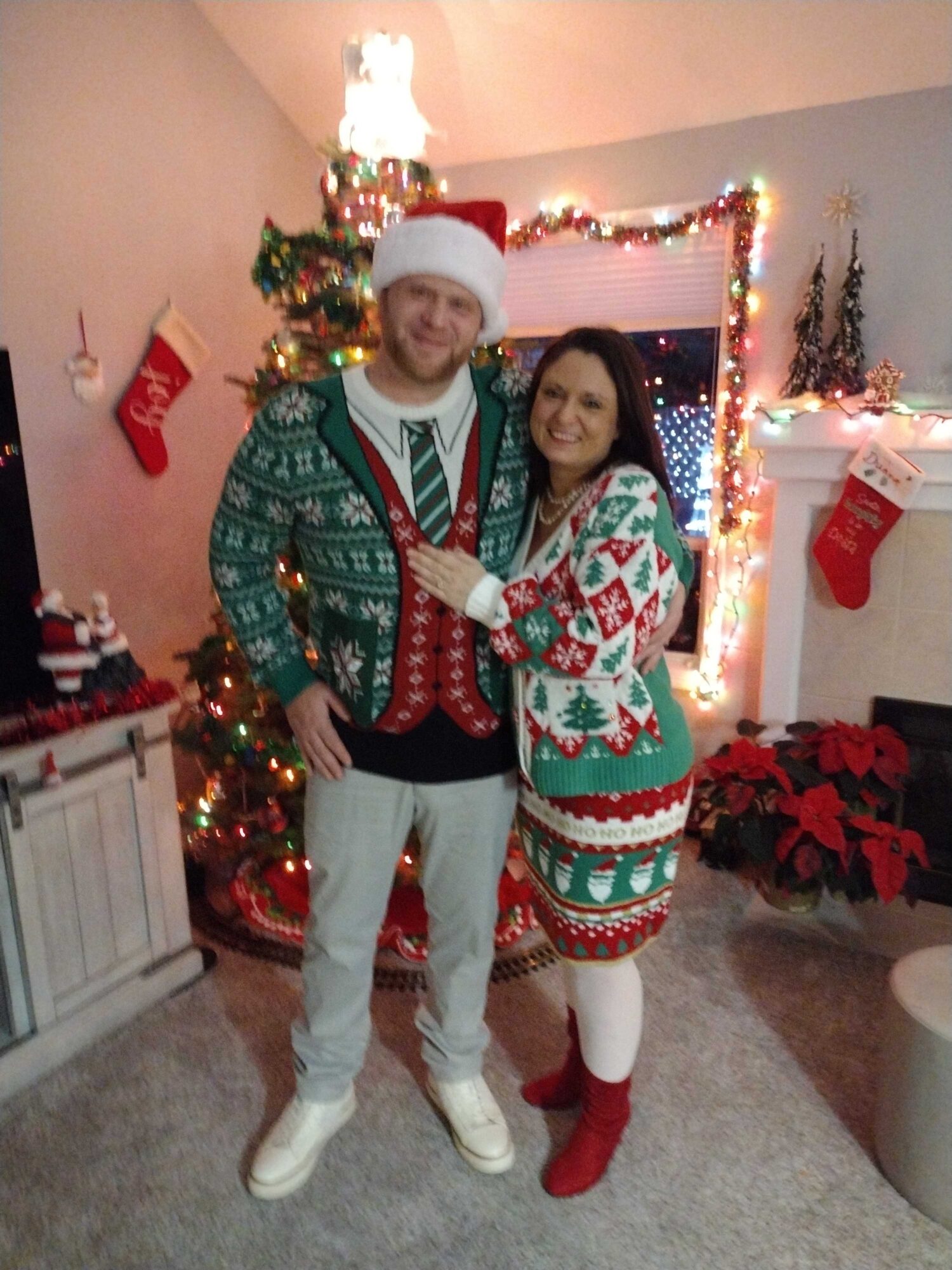 A smiling couple in Christmas sweaters and Santa hat stands in front of decorated Christmas tree and fireplace.