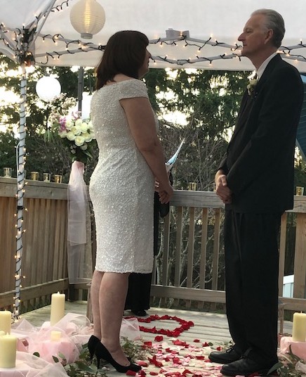 A woman in a white dress and black heels stands facing a man in a dark suit during a wedding ceremony outdoors, with decorated surroundings.