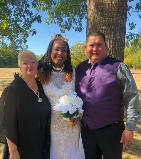 Three people standing outdoors near a tree, smiling, with a woman in a white dress holding a bouquet, and two others in dark and purple clothing.