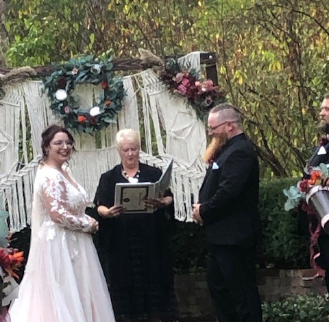 Wedding ceremony outdoors with bride, groom, officiant, and a person holding a book, decorated with flowers and greenery.
