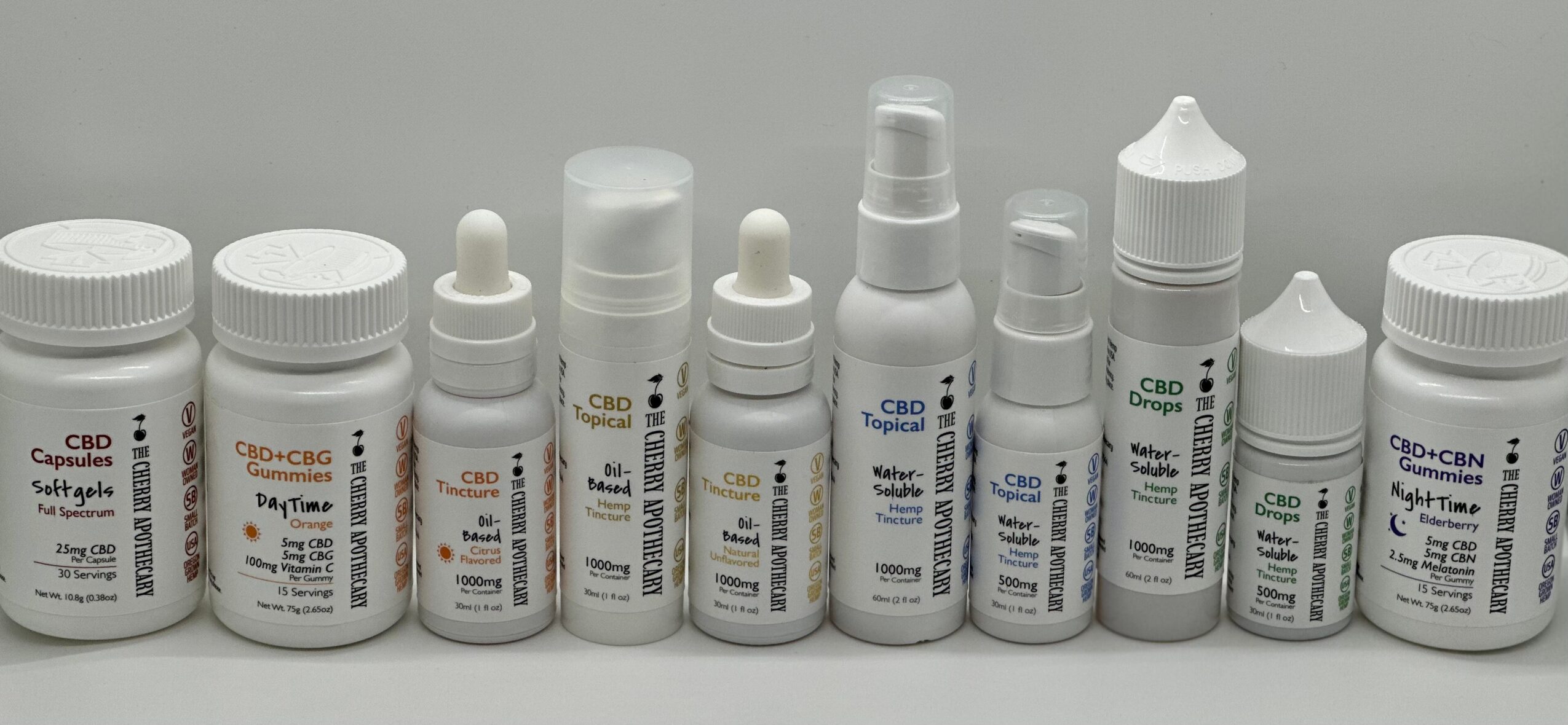 Line of various CBD product bottles with white caps and labels, some with droppers and spray nozzles, on a plain background.