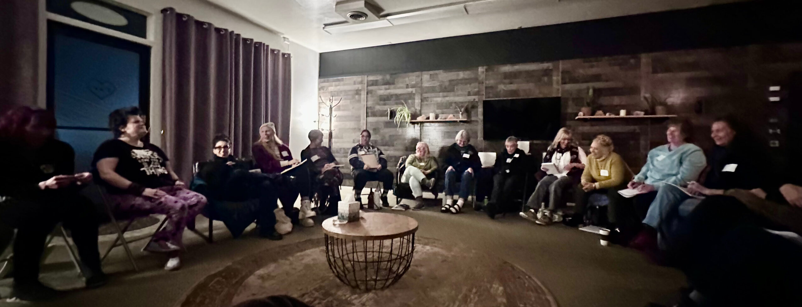 Group of people sitting in a circle in a dimly lit room with dark walls and curtains, some holding papers or notebooks.