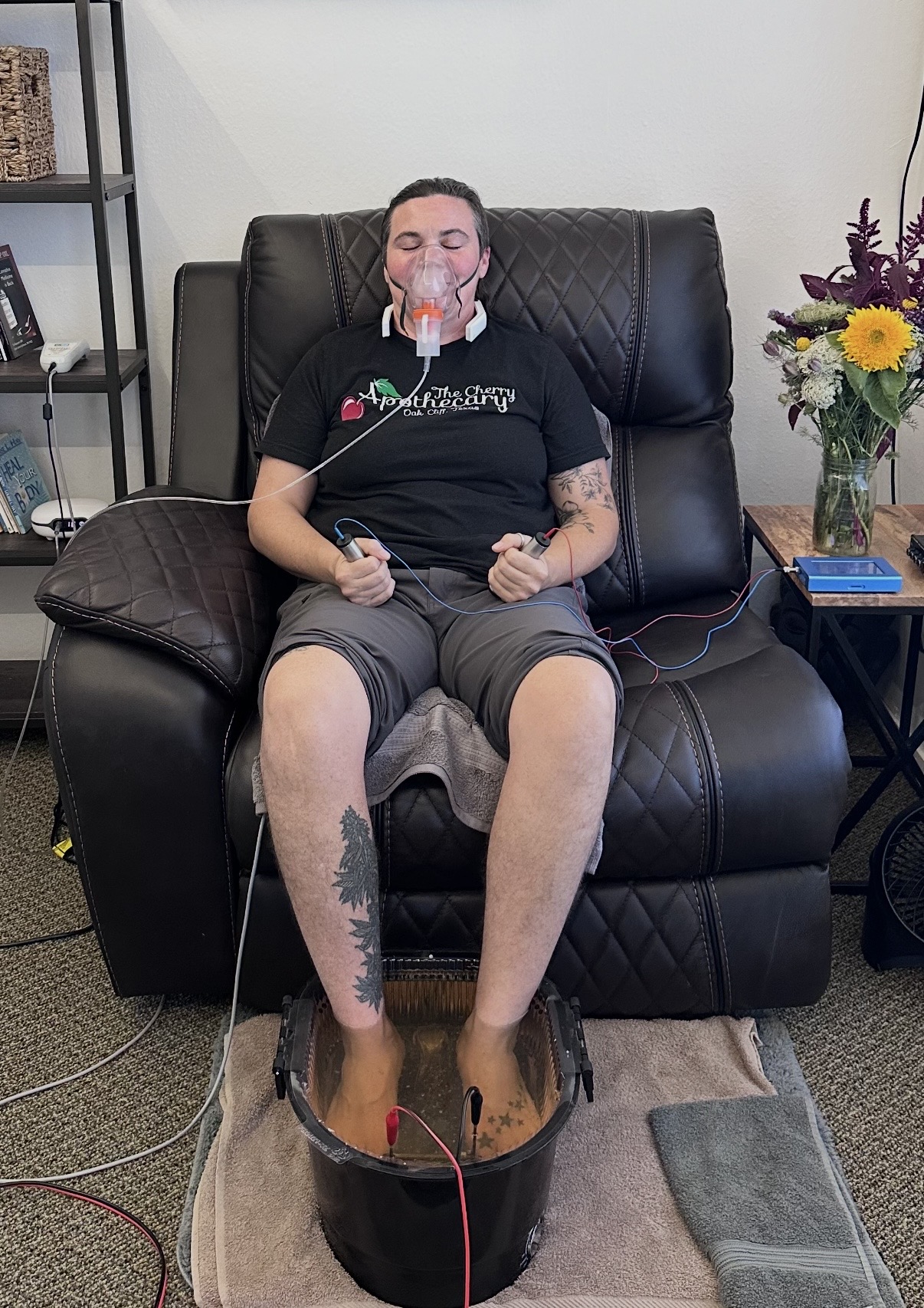 Person sitting on a black leather chair with feet in a wooden foot bath, holding a remote, with a flower arrangement nearby.