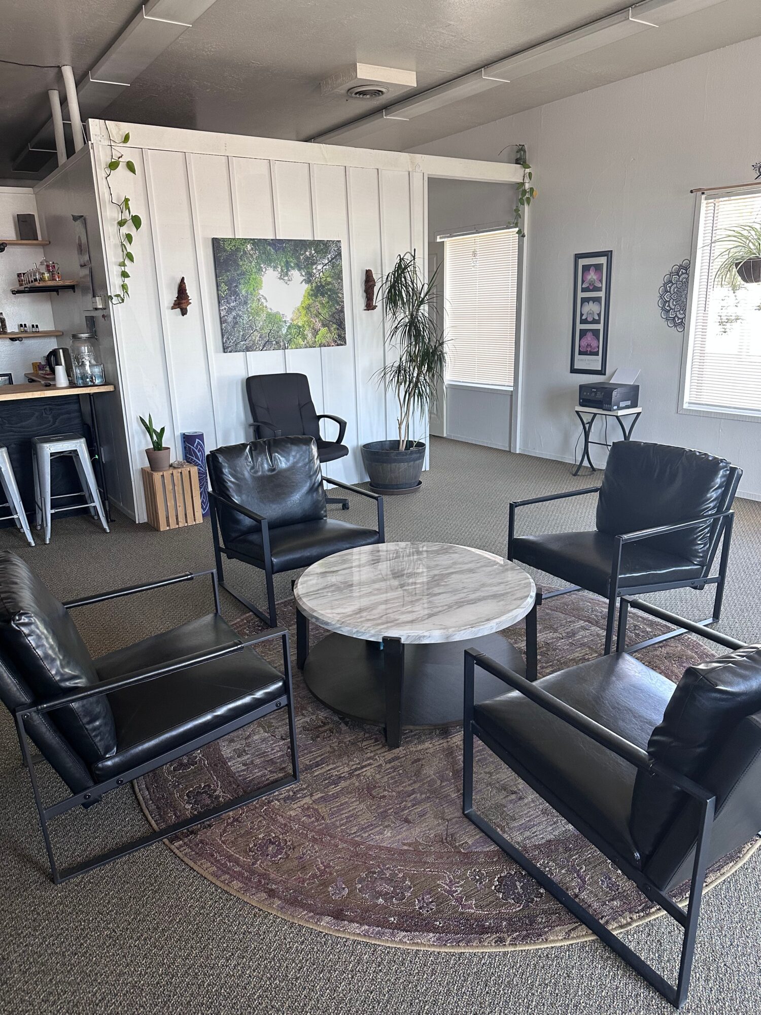 Four black chairs arranged around a round marble-topped table in a room with white walls, plants, and windows.