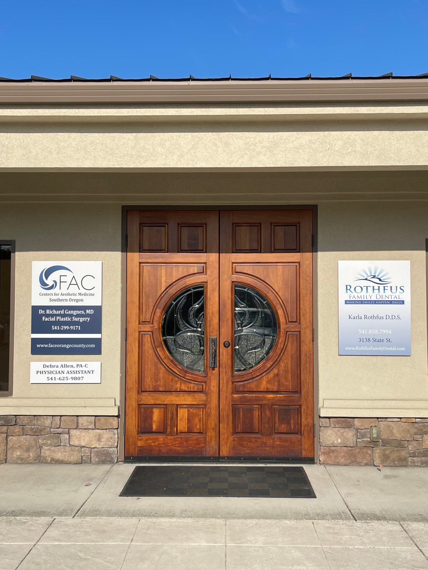 Wooden door with circular glass window, beige wall, and signs outside building, with sidewalk in front.