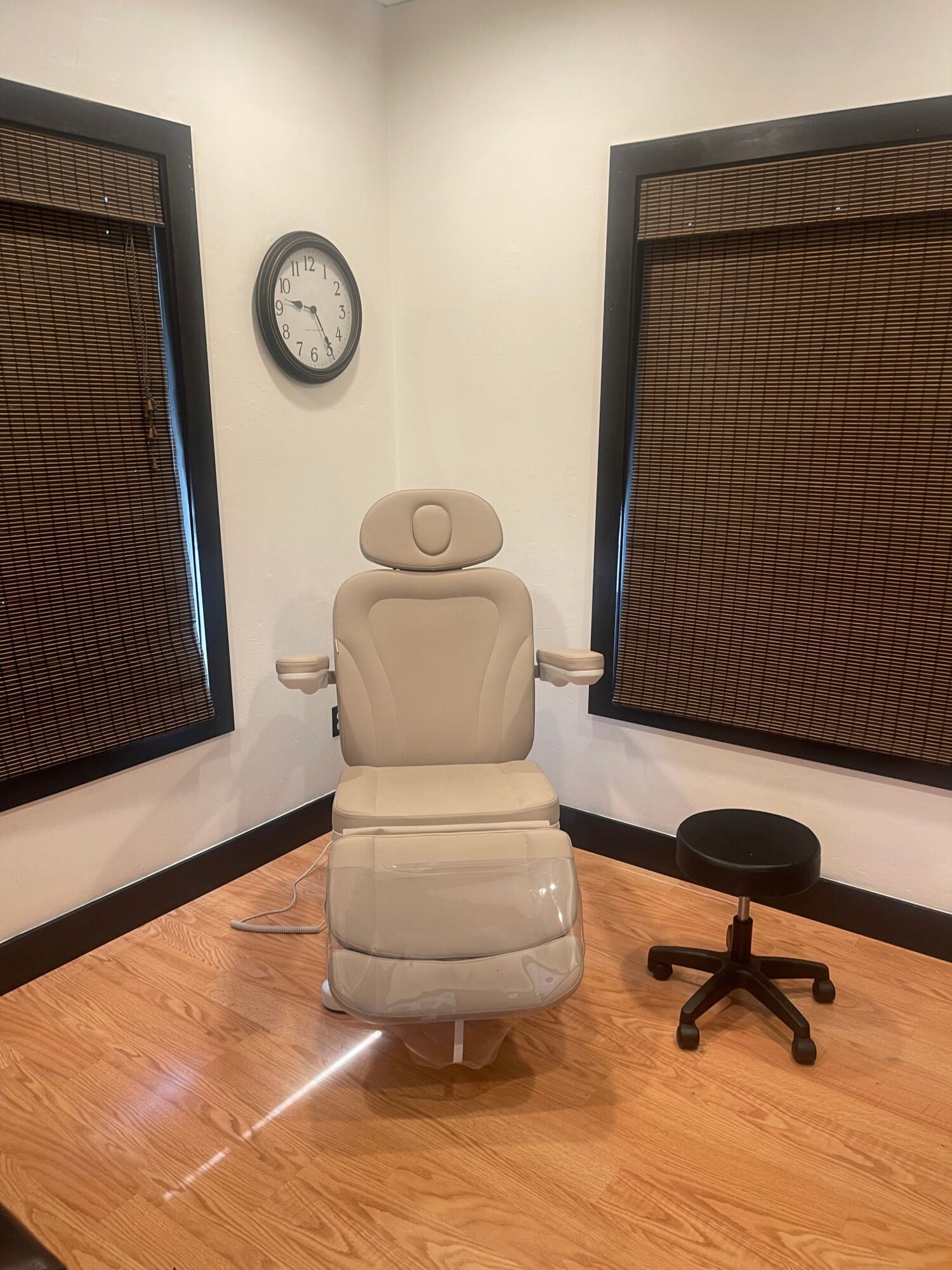 Reclining beige chair with armrests in a room with wooden floor, wall clock, and a black-framed window or screen.