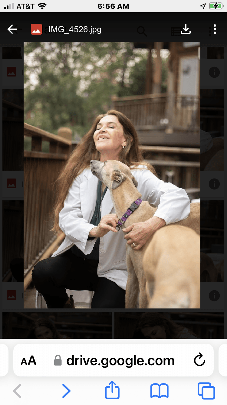 Woman smiling outdoors with a dog, holding a leash, in a natural setting with trees and a wooden structure behind.