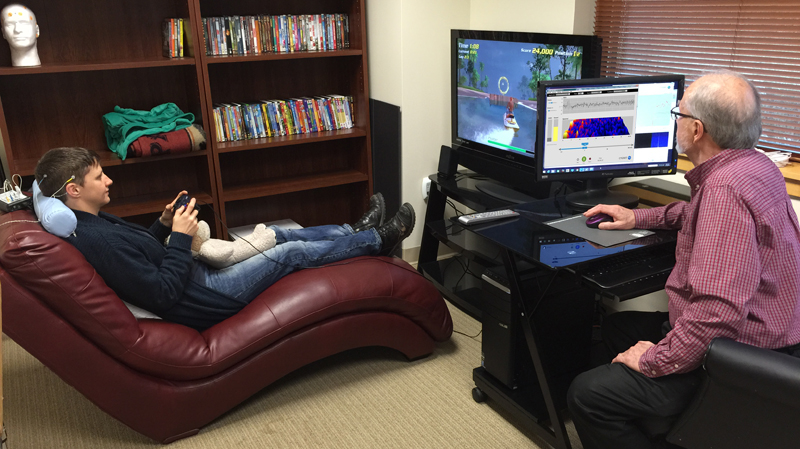 Young boy lying on a red couch playing video games, facing an older man at a computer station, in a room with bookshelves.