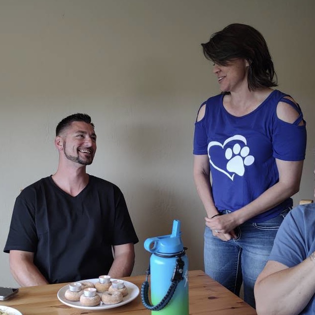 Two people smiling and talking at a table with a plate of cookies, one standing and one sitting, in a room.